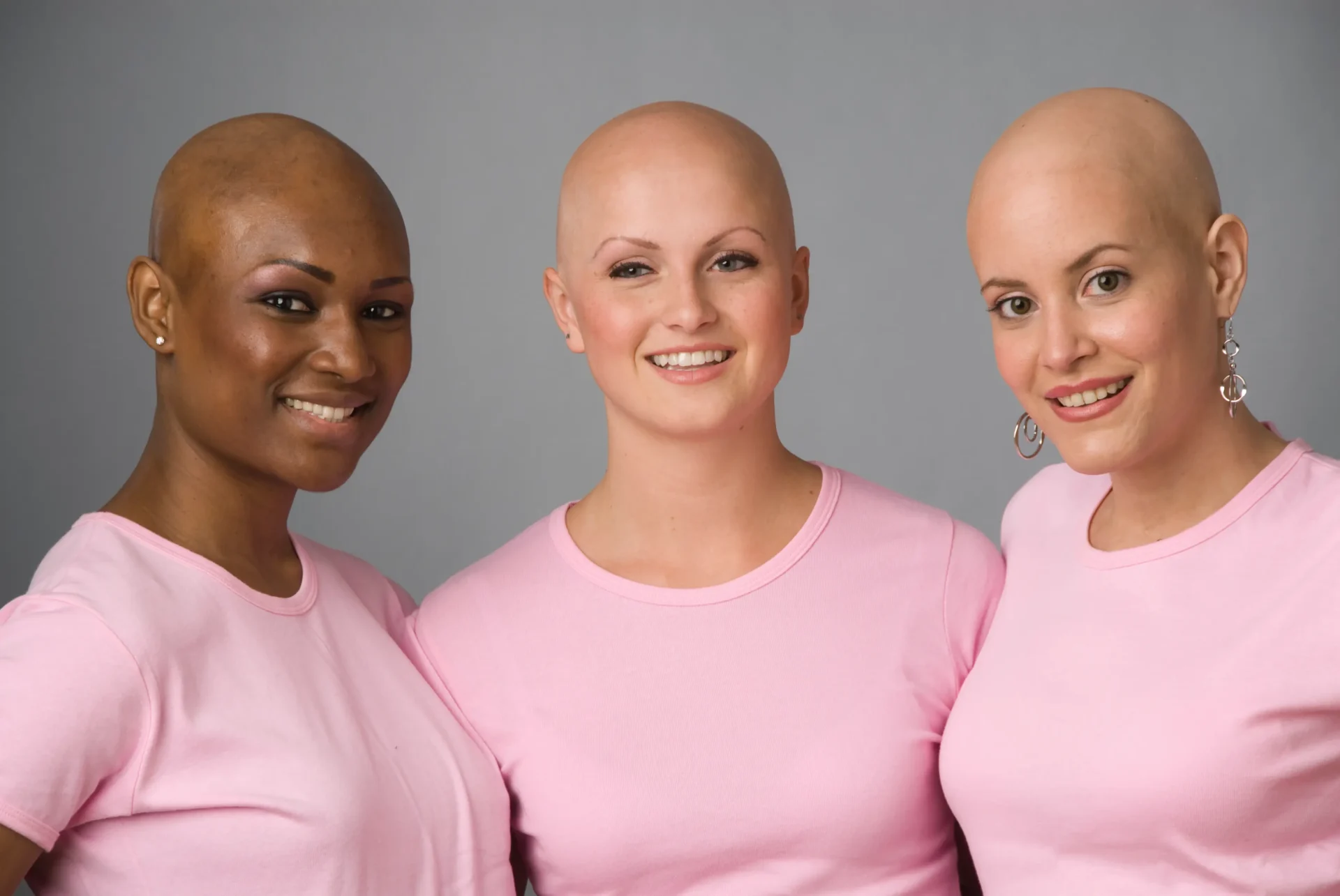 Three women standing together, wearing pink shirts with a gray background